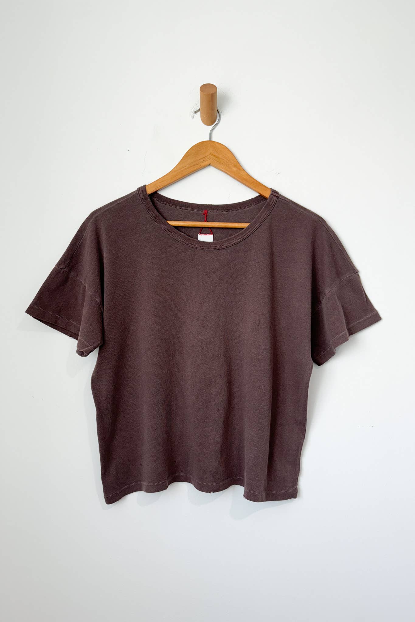 A chocolate brown, crew neck t-shirt hanging on a wooden hanger against a white wall.