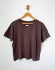 A chocolate brown, crew neck t-shirt hanging on a wooden hanger against a white wall.