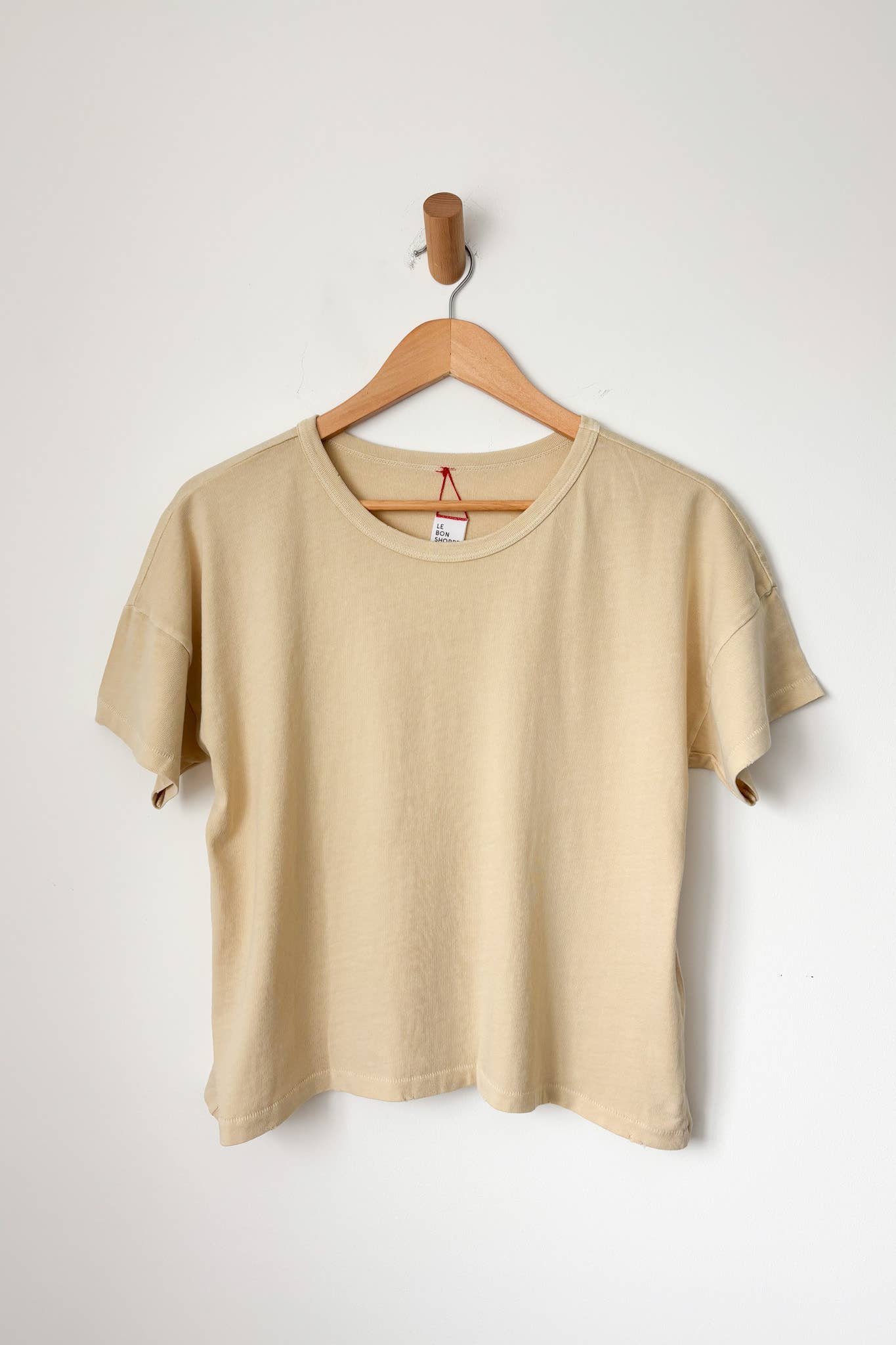 A butter beige t-shirt hanging on a wooden hanger against a white wall.
