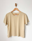 A butter beige t-shirt hanging on a wooden hanger against a white wall.